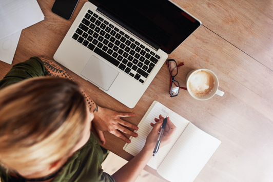 Overhead view of person writing in notebook next to laptop computer