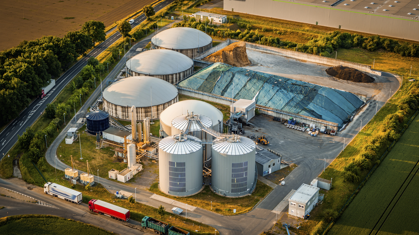Aerial view of a modern biogas facility in golden light, highlighting its large silos, covered feedstock storage, and circular fermentation tanks. Located near agricultural fields , the photo emphasizes the intersection of sustainable energy and rural infrastructure