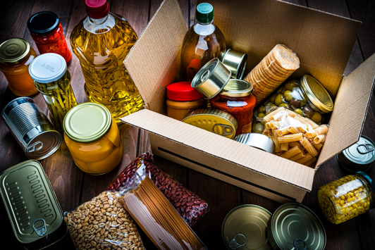Various foods in bottles cans tins and packaging in and around a box