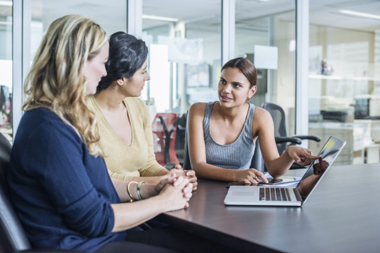 three people siting at a desk with a laptop talking