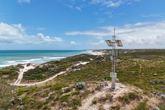 A tall Cel-fi repeater tower in the foreground with rolling hills and a coastline in the background.